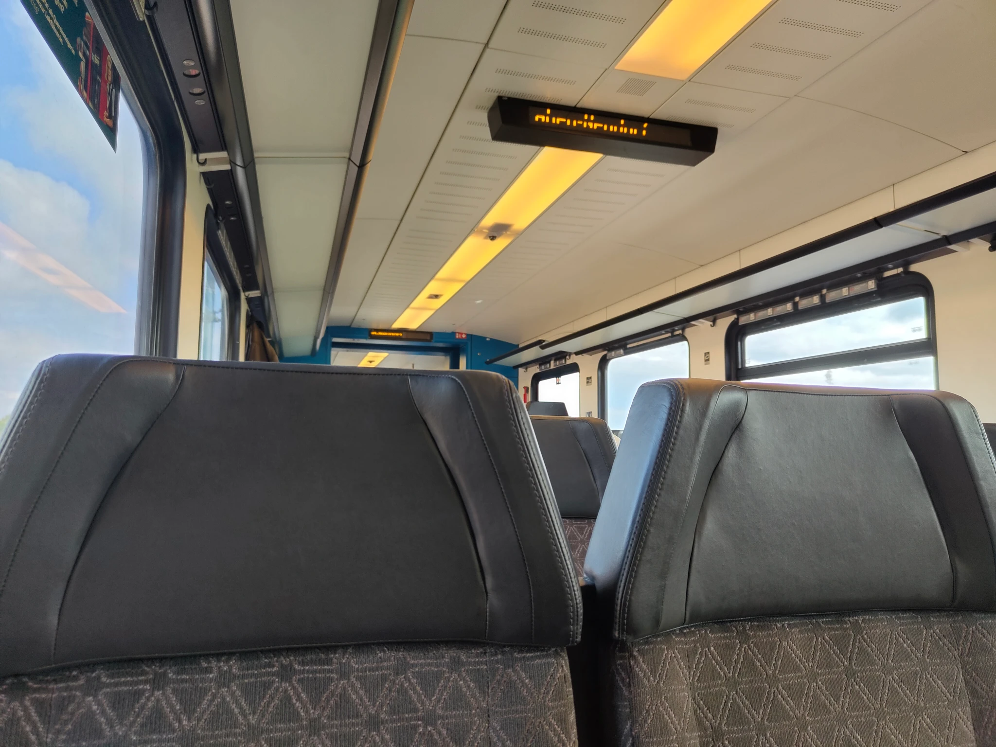 Photo of the interior of the train, taken in such a way that no people are visible, which means mostly the headrest and the ceiling are visible. The characteristic FLIRT 3 rectangular gangway between units can be visible in the background, in this train coloured blue. The seats have a triangular pattern on them.